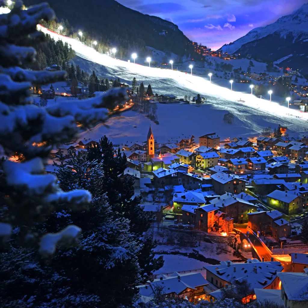 Veduta notturna di un villaggio alpino innevato con pista da sci illuminata.