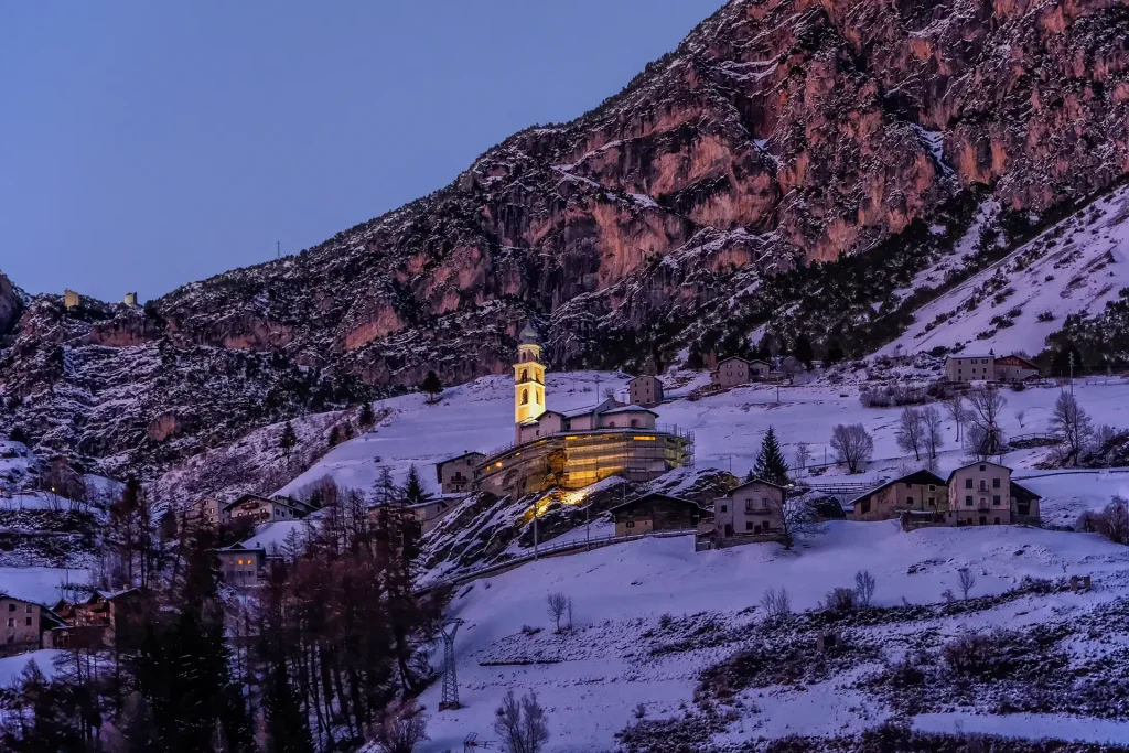 Paesaggio invernale di un villaggio alpino con una chiesa illuminata al tramonto.