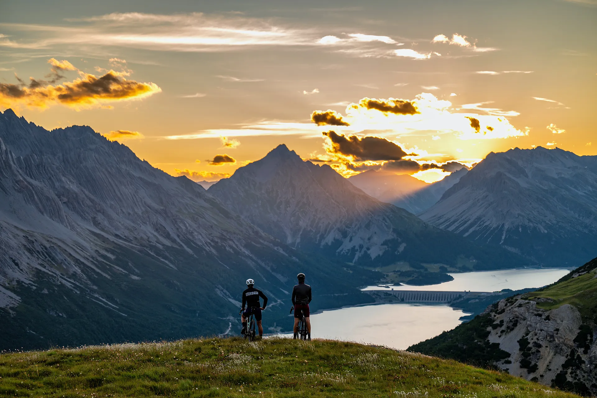 Due ciclisti ammirano il tramonto in montagna con vista su un lago.