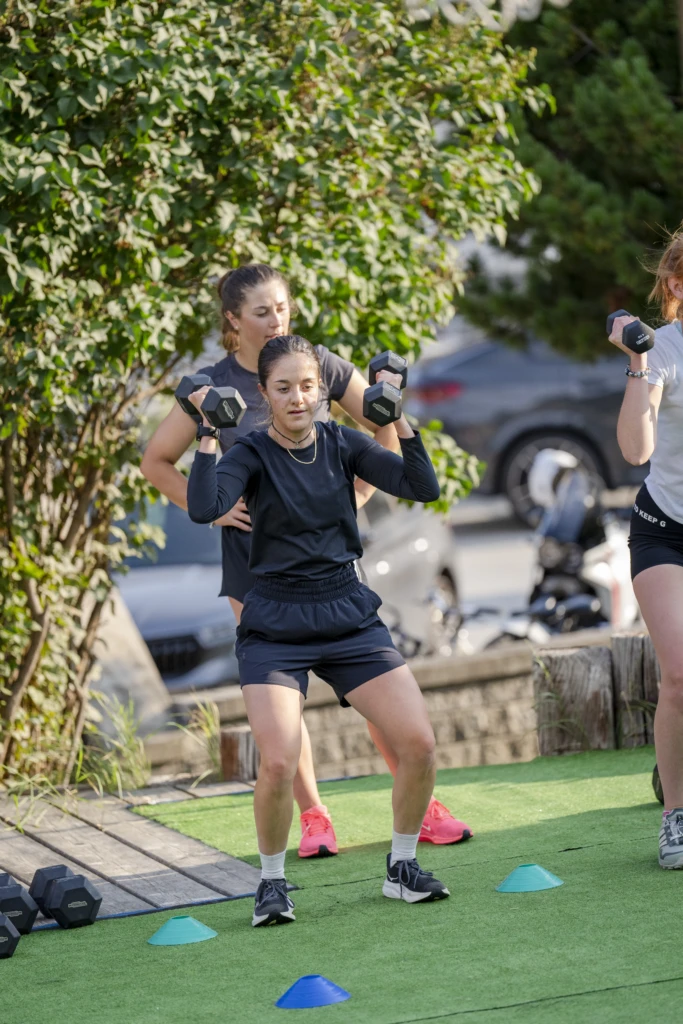 Three women exercising with dumbbells outdoors on a grassy area.