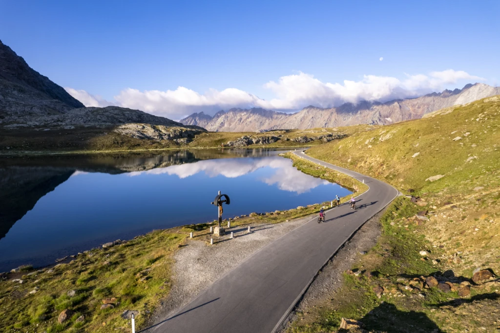 Cyclists ride on a scenic road beside a tranquil lake and mountain landscape.