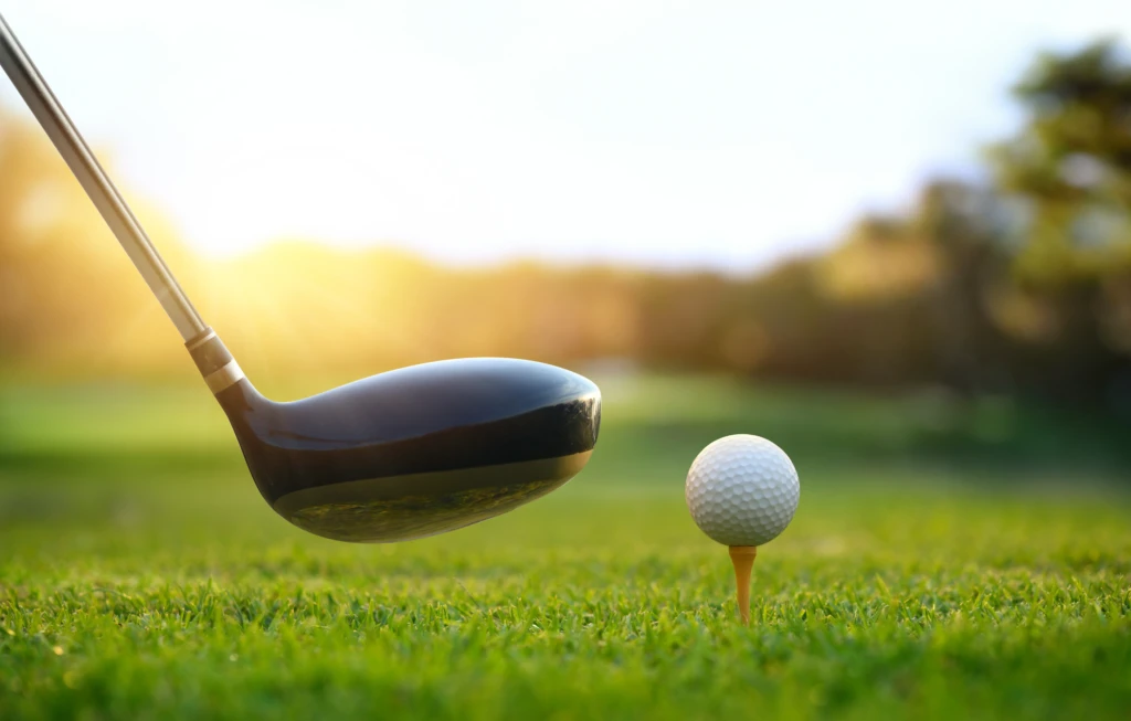 A golf club is about to hit a golf ball on a tee in the sunlight.