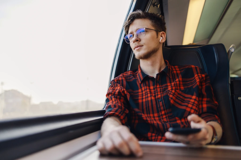 Giovane uomo con camicia a quadri rosso-nera guarda fuori dal finestrino di un treno, tenendo un cellulare.