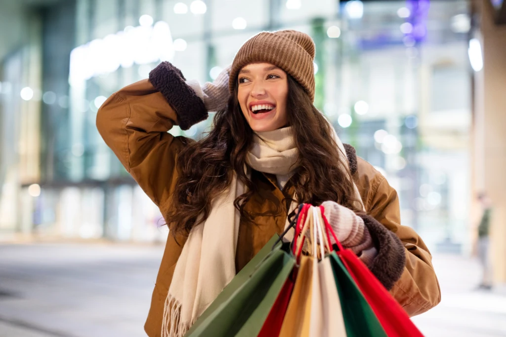 Donna sorridente con buste della spesa in abbigliamento invernale davanti a un centro commerciale.
