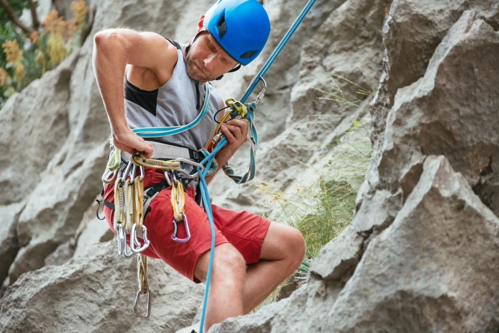 A climber wearing red shorts and a blue helmet adjusts their gear on a rocky surface.