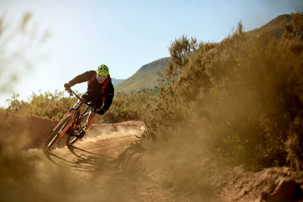 A cyclist rides a mountain bike on a dirt trail surrounded by bushes and hills.