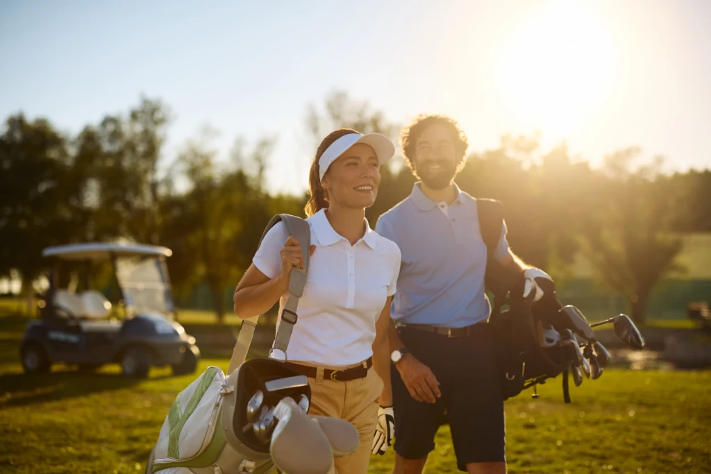 A man and woman carrying golf bags walk on a sunny golf course.
