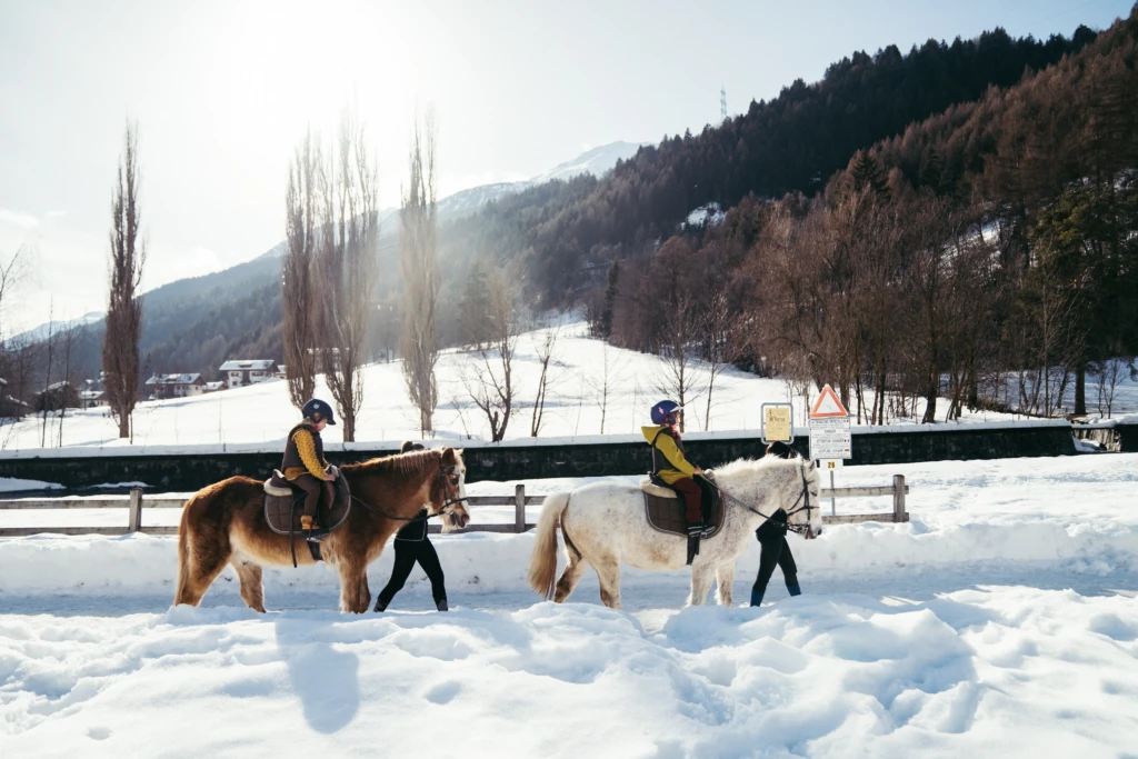 Due bambini a cavallo guidati da adulti su un sentiero innevato in montagna.