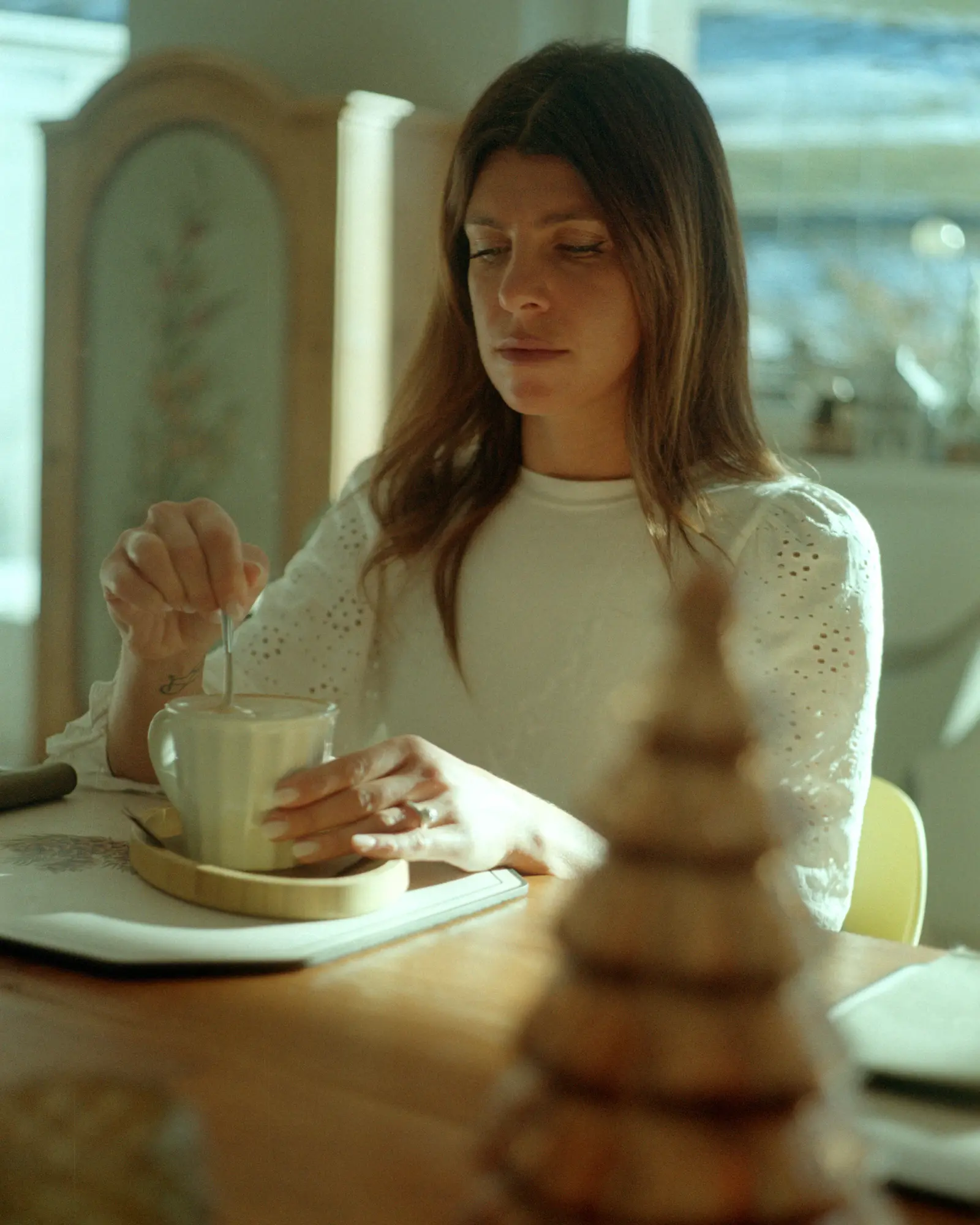 A woman stirs a hot drink at a sunlit table.