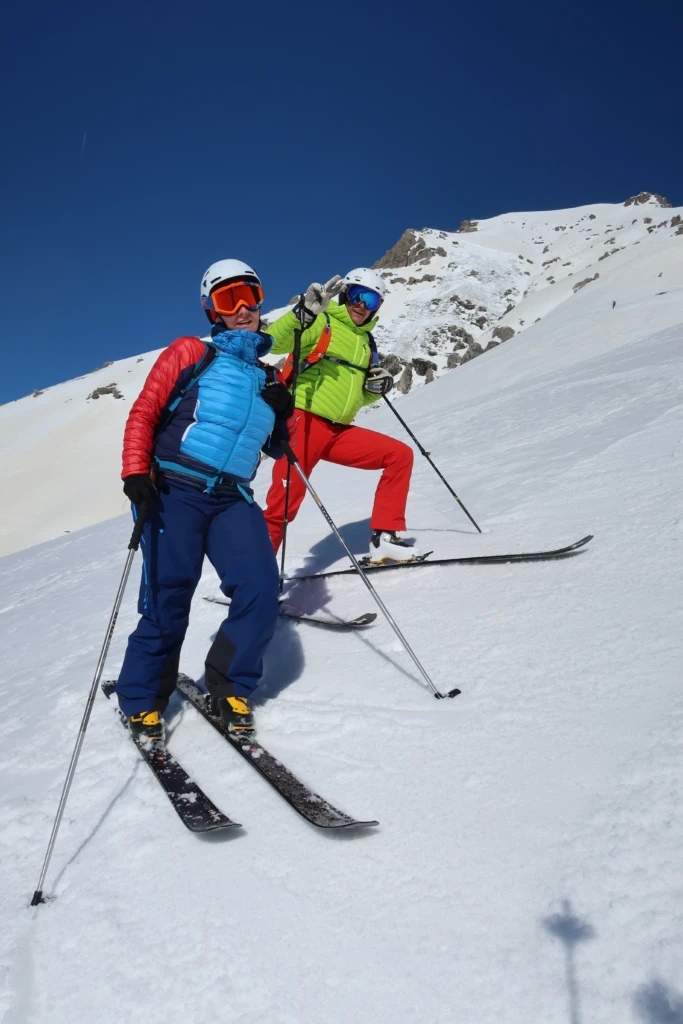 Due persone sorridenti sciano su una pista innevata sotto un cielo blu.