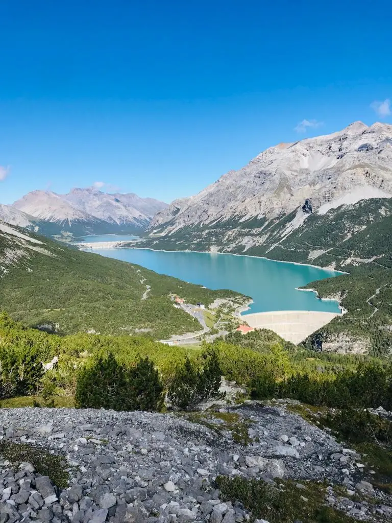 Veduta panoramica di un lago alpino circondato da montagne e foreste sotto un cielo azzurro.