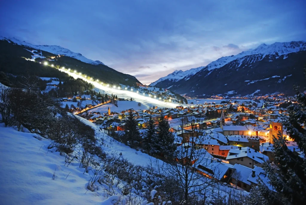 Paesaggio invernale serale di una cittadina alpina con pista da sci illuminata.