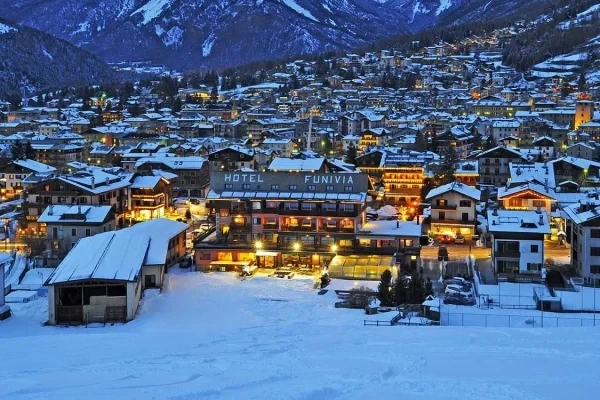 Vista notturna di una città montana innevata con un hotel illuminato al centro.