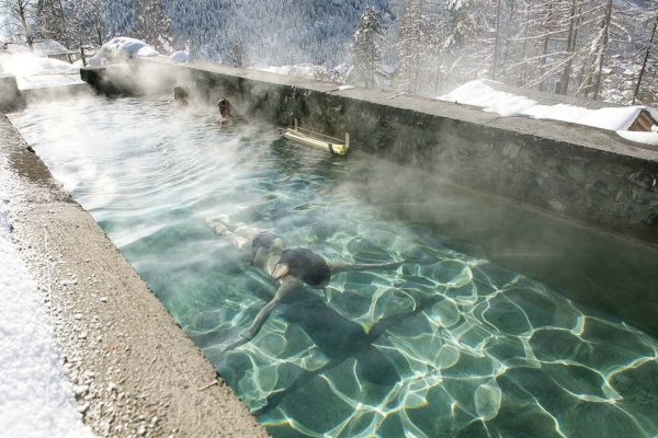 People relax in a steaming outdoor pool surrounded by snow-covered trees.